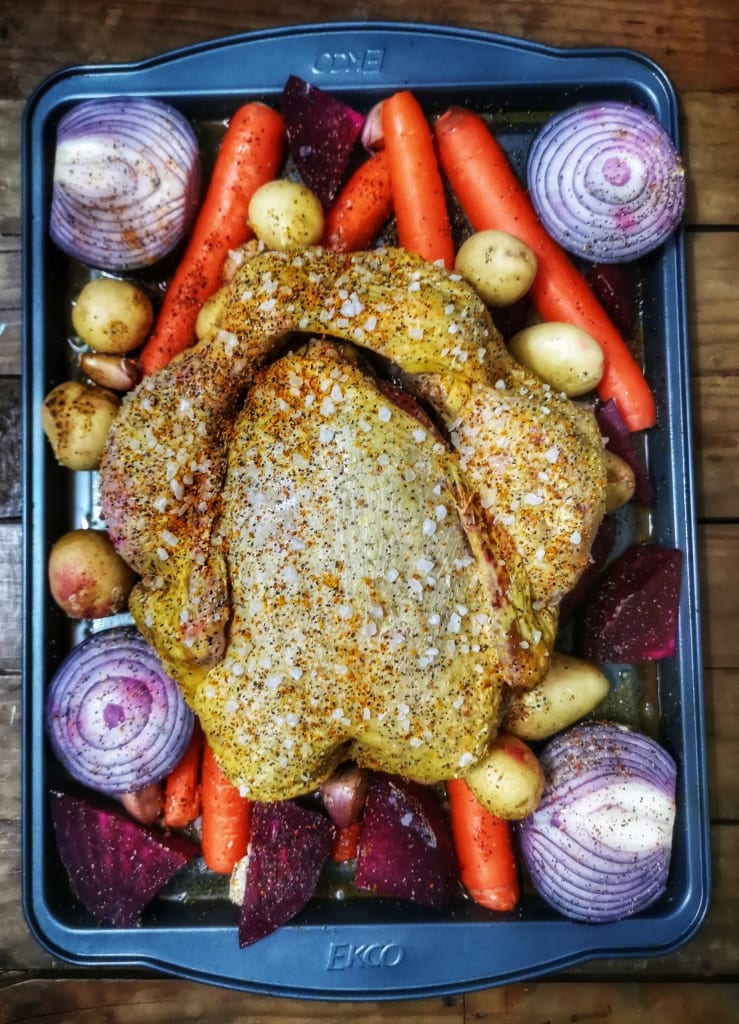 Full chicken on a tray with root vegetables underneath, before it's roasted in an oven.